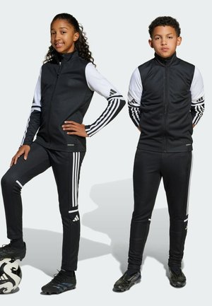 Survêtements de sport noirs avec des accents blancs et trois bandes sur les manches et les pantalons. Modèle portant des chaussures de football noires, tenant un ballon de football.