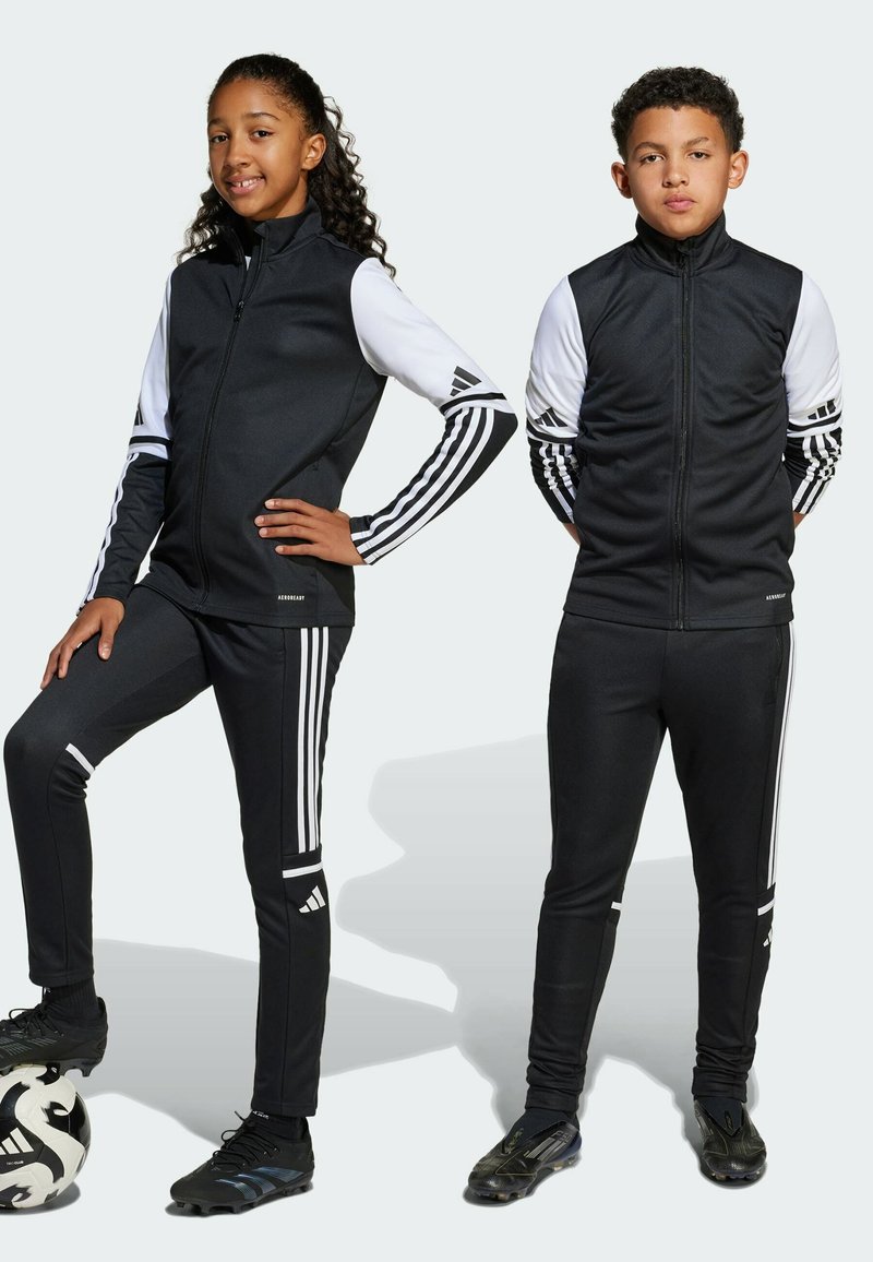 Czarne dresy sportowe z białymi akcentami i trzema paskami na rękawach oraz nogawkach. Model w czarnych butach piłkarskich, trzymający piłkę nożną.