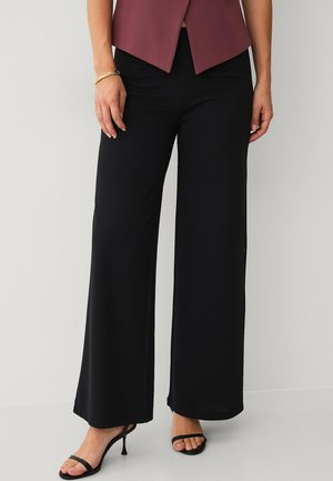 Kvinde iført sorte bukser med vide ben, bordeaux top, guldbøjle og sorte højhælede sandaler, stående op ad en hvid væg.