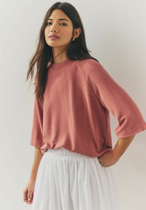 Pulover supradimensionat de culoare roz cu mâneci scurte și guler ondulat, asortat cu o fustă albă, plisată. Textură netedă, potrivire casuală.