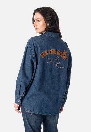 Kvinde med mørkt hår iført en blå denimjakke og jeans, jakken viser broderet tekst på ryggen "See the good in all things."