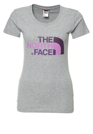 Kobieta w szarej koszulce The North Face, granatowych szortach do kolan i brązowych butach turystycznych, stojąca z jedną ręką w kieszeni.