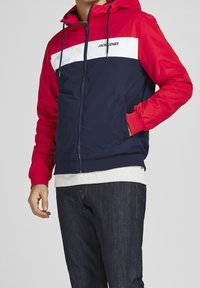 Námořnická a červená prošívaná bunda s bílým horizontálním pruhem, má přední zip, kapuci a boční kapsy. Na pruhu je nápis "JACK&JONES".