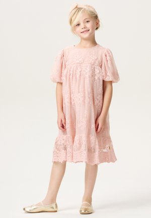 Roze jurk van kant met bloemenbroderie, pofmouwen en een gerimpelde zoom. Gepaard met gouden ballerina's en een bijpassende haarband.
