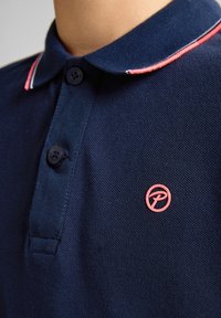 Námořnická modrá polo košile s texturovanou tkaninou; má tříknoflíkovou légu, kontrastní červené a bílé pruhy na límci a růžové logo.