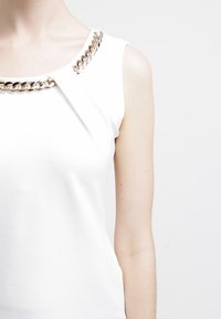 Bílý top bez rukávů s texturovaným povrchem, zdobený zlatým řetízkem u výstřihu a s propracovaným designem ramen.