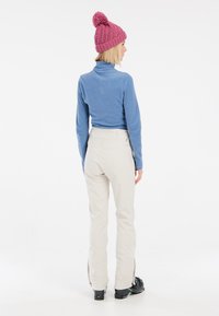 Maglione a maniche lunghe in fleece blu e pantaloni da sci color crema. Il modello indossa un berretto di maglia rosa con pom-pom e scarponi da sci neri con dettagli.