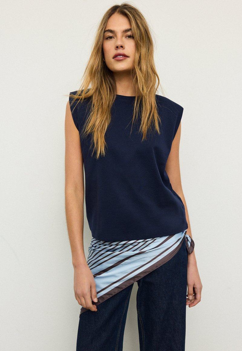 Žena s dugom plavom kosom koja nosi tamnoplavu majicu bez rukava i tamne hlače, s plavim i smeđim prugastim šalom vezanim oko struka.