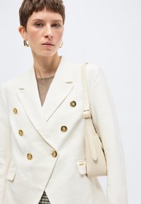 Γυναίκα με κοντά μαλλιά φοράει λευκό σακάκι διπλής σειράς με χρυσά κουμπιά, κρατώντας μια κρεμ τσάντα ώμου, σε απλό φόντο.
