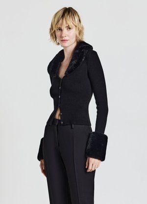 Donna che indossa un cardigan nero a coste con collo e polsini in pelliccia, abbinato a pantaloni neri su misura, in piedi davanti a uno sfondo chiaro e uniforme.