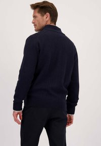 Man met zijn rug naar de camera, gekleed in een donkerblauwe trui over een geblokte shirt en donkere broek, tegen een effen lichte achtergrond.