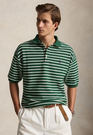 Joven con camisa polo de rayas verdes y blancas, pantalones crema y cinturón trenzado marrón, manos en los bolsillos, mirando hacia la izquierda.