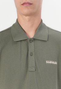 Oliven grønn polo skjorte laget av teksturert bomull, med kragehals, to knapper foran og en logo på venstre brystområde.