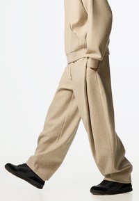 Bež široke trenirke od mekanog tkanja s plisiranim dizajnom, bočnih džepova i elastičnim pojasom, uparene s crnim tenisicama.