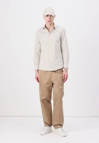 Stripet button-up skjorte i lys beige, kombinert med brune bukser. Tilbehør inkluderer en beige caps og hvite joggesko. Casual passform.