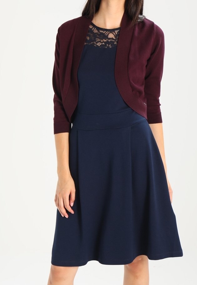 Kvinna i en marinblå knälång klänning med spetsdetaljer vid halslinningen och en kort ärmad burgundy cardigan med trekvartslånga ärmar.