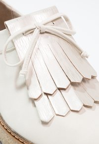 Gros plan d'un mocassin en cuir blanc avec un détail en cuir frangé métallique et un lacet noué sur le dessus.