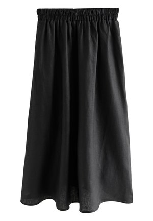 Młoda kobieta ubrana w czarną bezrękawnikową, zapinaną na guziki bluzkę i szerokie czarne spodnie stoi na drewnianym chodniku wśród dużych skał.