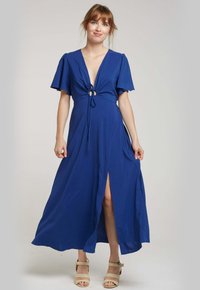 Robe maxi bleue avec un décolleté en V profond, des manches courtes à volants, un détail noué à l'avant et une fente sur le côté. Tissu doux. Sandales simples beiges portées.