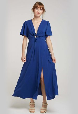 Robe maxi bleue avec un décolleté en V profond, des manches courtes à volants, un détail noué à l'avant et une fente sur le côté. Tissu doux. Sandales simples beiges portées.