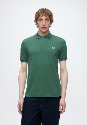 Joven con cabello castaño claro, vestido con una camiseta polo verde de manga corta y vaqueros azul oscuro, de pie frente a un fondo blanco liso.