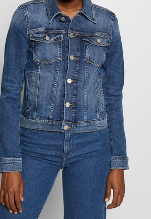 Persoon draagt een getailleerd blauw spijkerjasje dichtgeknoopt met bijpassende blauwe spijkerbroeken tegen een effen achtergrond.