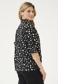 Schwarze Bluse mit weißen abstrakten Mustern, kurzen Puffärmeln und einem Stehkragen. Weicher Stoff mit lockerer Passform.
