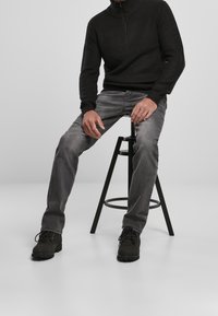 Homme en pull en maille noir, jean gris et bottes noires, assis sur un tabouret en métal noir devant un fond gris uni.