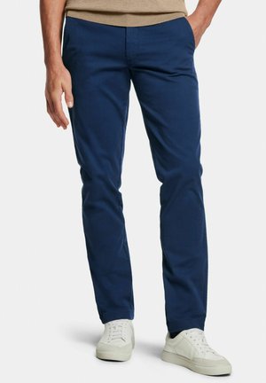 Homme portant un pantalon bleu marine ajusté et des baskets blanches, debout avec une main dans la poche devant un fond uni.