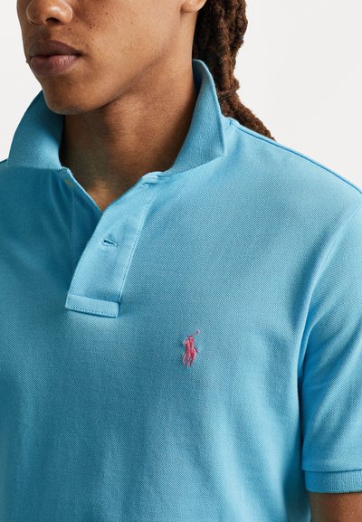 Tricou polo de culoare albastru deschis, realizat dintr-un material texturat, având un guler cu două butoane și un mic logo roz brodat pe piept.