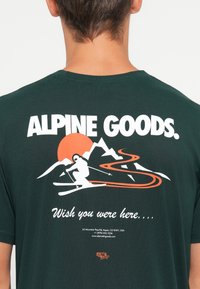 Baksiden av en mørkegrønn skjorte med hvitt og oransje grafikk som viser en skikjører, en svingete fjellsti, solnedgang og teksten "ALPINE GOODS. Ønsker du var her."