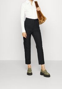 Svarta skräddarsydda byxor, strukturerad vit skjorta och olivgröna slip-ons. Tillbehören omfattar en brun pälsväska, vilket skapar en genomtänkt outfit.