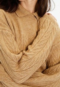 Suéter beige de punto trenzado con cuello acanalado y puños de canalé, que presenta un patrón texturizado en toda la prenda. Material suave y cálido.