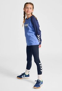Blå långärmad tröja med marinblå ärmar och vit logotyp; marinblå leggings med vit chevronmönster; blå sneakers med gröna accenter.