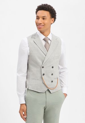 Junger Mann, der eine graue, doppelreihige Weste mit einer Kette, ein weißes Hemd, eine beige Krawatte und hellgrüne Hose trägt, lächelt und seitlich schaut.