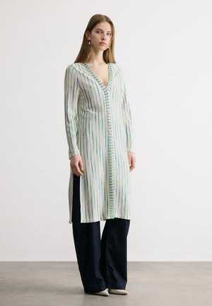 Fiatal nő hosszú, csíkos, gombos kardigánt visel fekete, széles szárú nadrág fölött, bézs hegyes orrú cipőben, egyszínű háttér előtt állva.