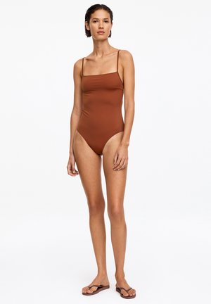 Kvinde stående iført en brun heldragt med tynde stropper og brune flip-flops på en hvid baggrund.