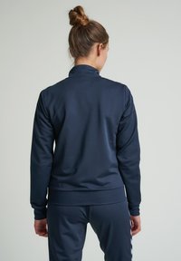 Marinblå idrottsjacka med hög krage, slät textur och figurnära silhuett. Har långa ärmar och ribbad fåll, i kombination med matchande byxor.