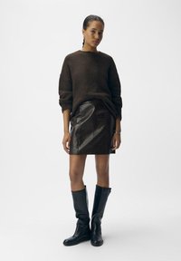 Maglione lavorato a maglia marrone, gonna in pelle nera con motivo coccodrillo e stivali alti fino al ginocchio in pelle nera. La modella è in piedi contro uno sfondo neutro.
