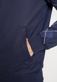 Chaqueta de color azul marino con cremallera, fabricada en un tejido texturizado, que cuenta con un bolsillo lateral y líneas geométricas blancas en contraste en las mangas.