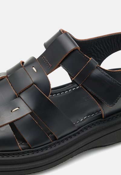 Sandale en cuir noir avec un design de lanière tissée, des coutures contrastées en tan et une semelle en caoutchouc robuste. Présente des accents en ton argenté et un dos ouvert.