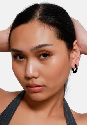 Orecchini a cerchio neri, design rotondo, texture liscia, indossati all'orecchio destro. La modella presenta capelli lisci e scuri su uno sfondo neutro.