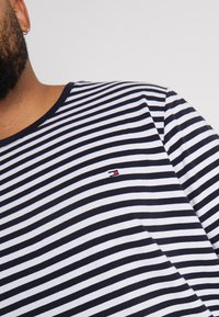 Vousatý člověk v námořnicky modrém a bílém pruhovaném tričku s malým červeným, bílým a modrým logem na hrudi, viditelný zlatý kroužkový náušnice.