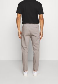 Lichtgrijze, op maat gemaakte broek van gladde stof, met twee achterzakken en taps toelopende pijpen. Gecombineerd met een zwart shirt en witte sneakers.