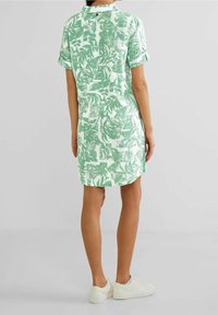 Sukienka z krótkim rękawem w jasnym zielonym kolorze z białym wzorem liści, z kołnierzykiem i guzikami z tyłu, zestawiona z białymi trampkami.