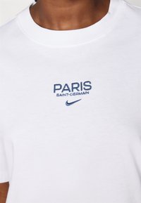 Valkoinen puuvillapaita, jossa on sininen brodeeraus: "PARIS SAINT-GERMAIN" pienen Nike-logon yläpuolella, pyöreä pääntie ja lyhyet hihat.