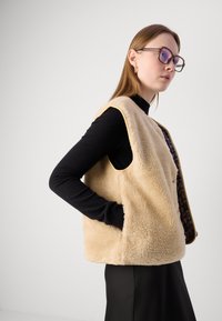 Beige pälsväst med ärmlös design och sidofickor, i kombination med en svart långärmad topp. Utmärkande mjuk textur och avslappnad passform.