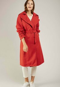 Manteau trench rouge avec une taille ceinturée, un devant double boutonnage, un large col et des manches retroussées. Porté sur un pantalon blanc et des baskets.