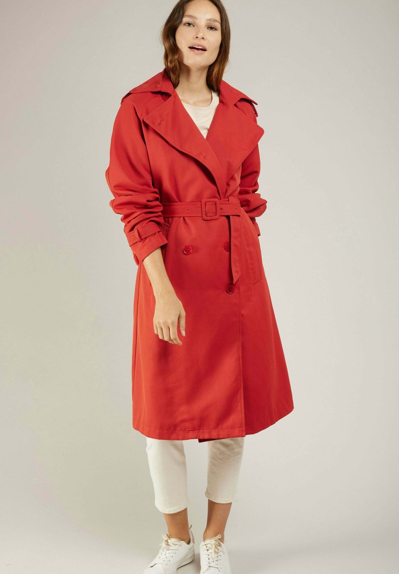 Manteau trench rouge avec une taille ceinturée, un devant double boutonnage, un large col et des manches retroussées. Porté sur un pantalon blanc et des baskets.