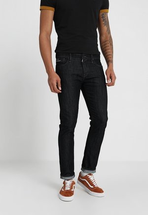 Jeans de mezclilla negra con un ajuste ceñido, que cuentan con cinco bolsillos y trabillas para cinturón, combinados con zapatillas de ante marrón y una camisa negra con ribete amarillo.